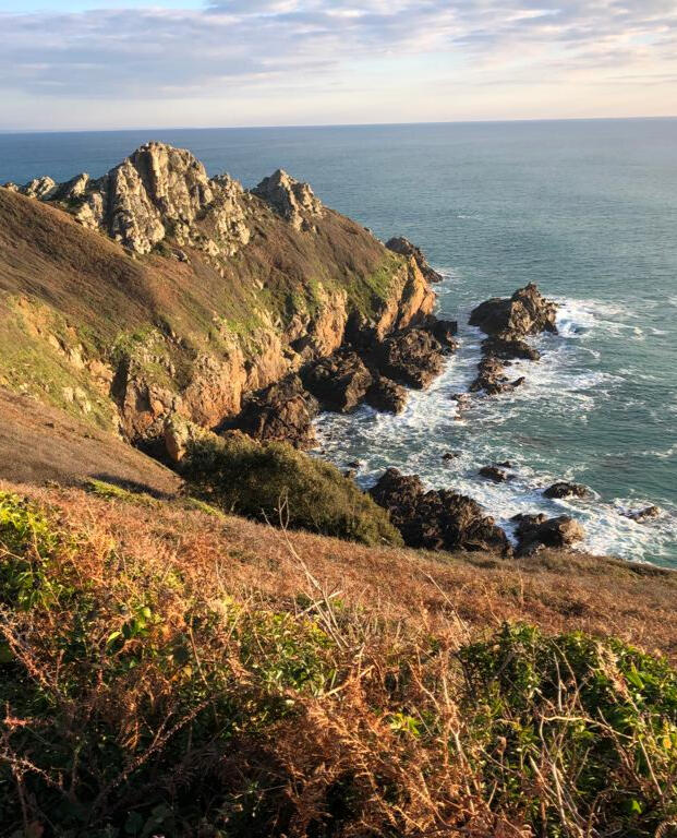 Photo taken in Cork on the south coast of Ireland. Brown granite cliffs glow in golden sunlight, lightly covered in grass and browning gorse. The sea is far below, frothing white on the rocks, and looks rather cold and cruel from up here.