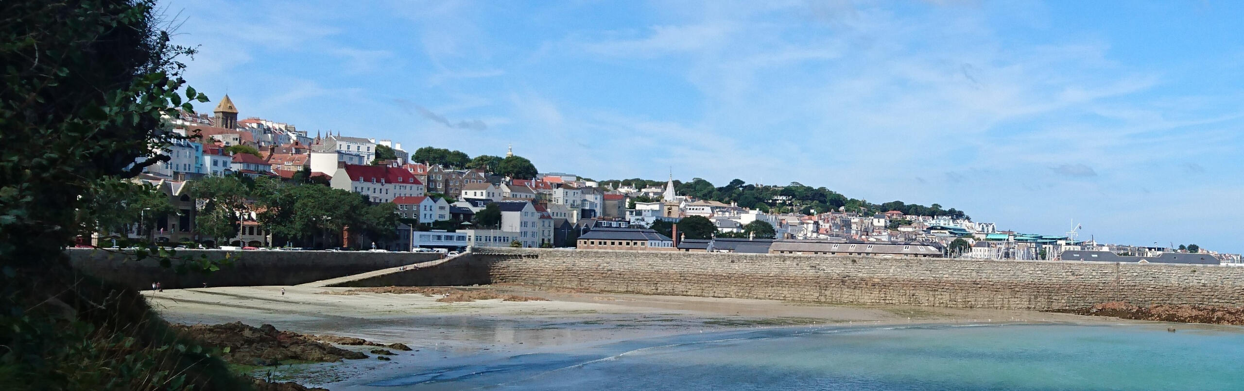 A photo of the seaside town of St Peter Port in Guernsey rises roof upon roof up the ridge amid dark green trees. There is a long granite pier, perpendicular to the town, leading to the castle out in the bay. The tide is out and is very blue.