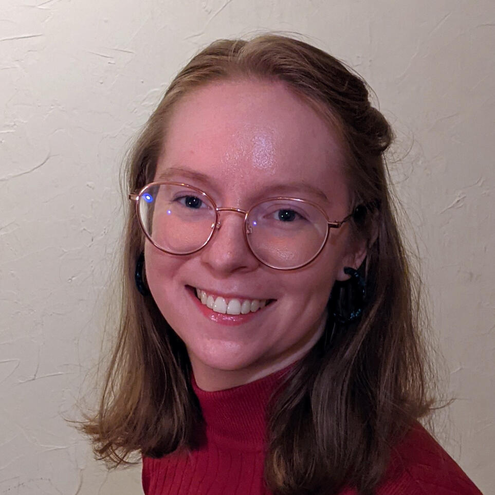 Headshot of Lillian. They are a white British person with mid-length auburn hair and round, pink-framed glasses.